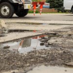 Potholes filled with water on a damaged road near construction cones.