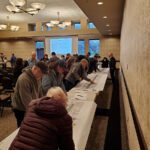 People are engaged in filling out forms at a long table in a room with large windows.
